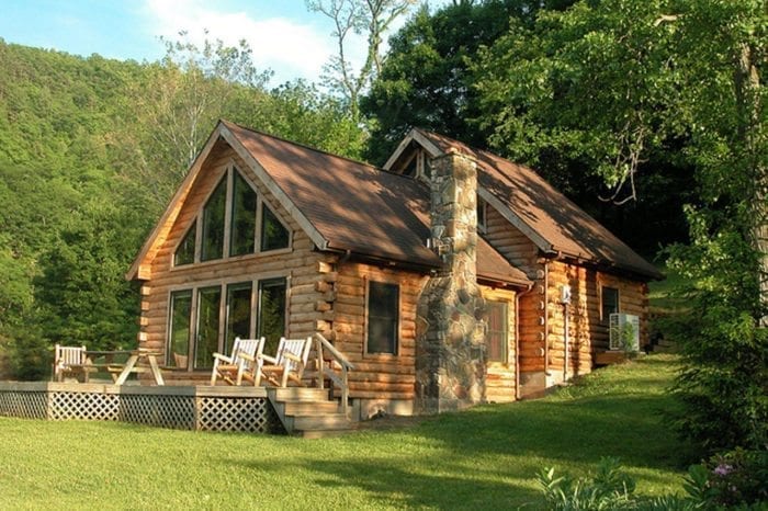 Exterior view of log cabin with large windows, outdoor deck, and stone chimney